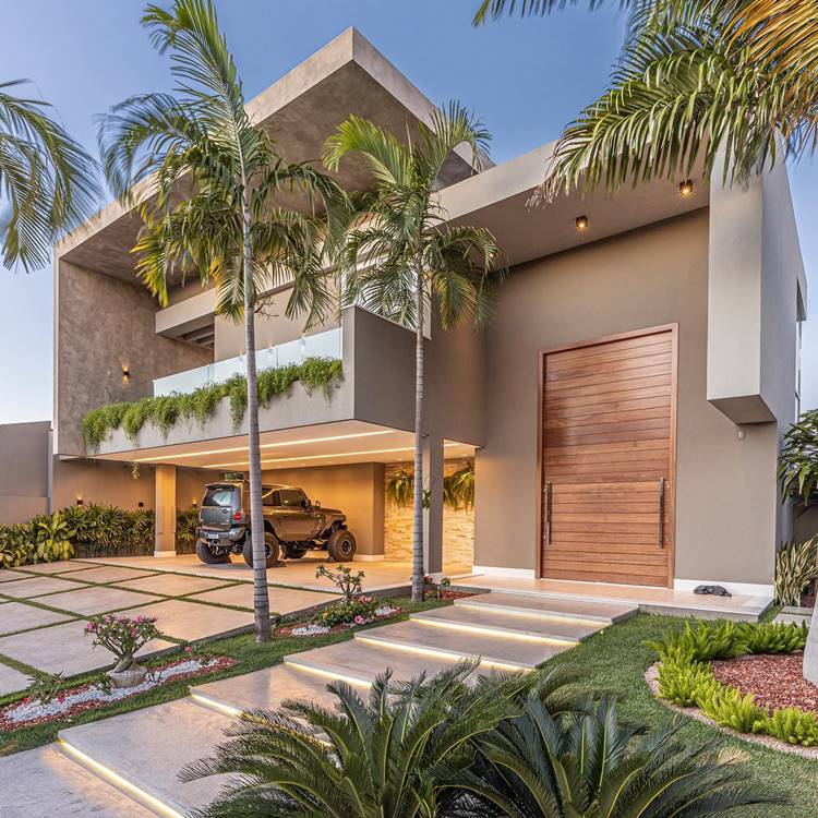 Fachada de casa moderna de dois andares durante o dia. A casa tem um design arquitetônico geométrico e uma cor neutra. Há uma porta de madeira na frente e um carro estacionado na garagem. Várias palmeiras e outras plantas decoram o jardim. O céu está claro e sem nuvens.