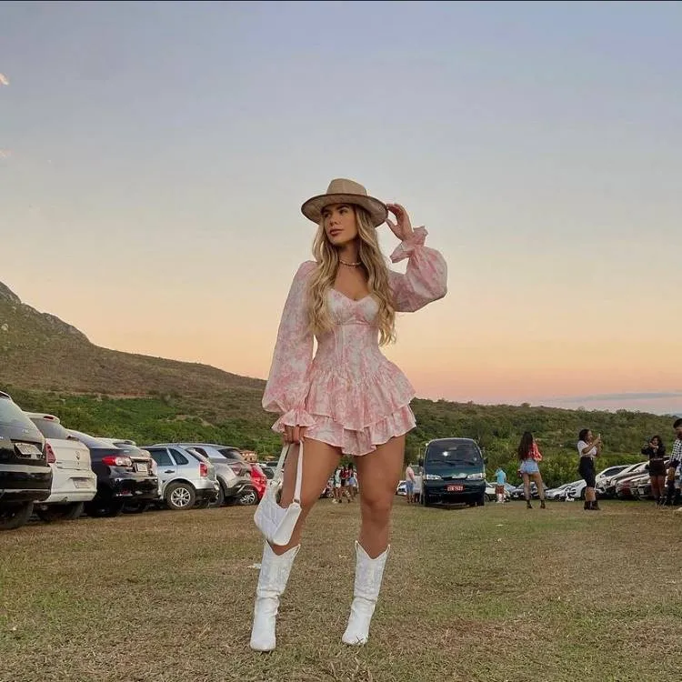 A imagem retrata uma jovem elegante em um evento ao ar livre ao pôr do sol. Ela usa vestido floral rosa claro, botas brancas e chapéu bege. Ao fundo, carros estacionados, pessoas conversando e colinas verdes sob um céu colorido.

