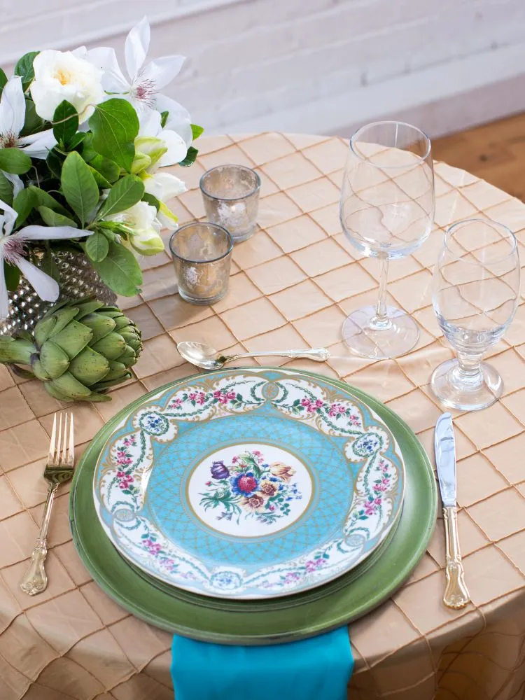 A imagem mostra uma mesa coberta com um pano de quadriculado bege. A mesa está servida com um prato verde abaixo de outro prato com um desenho floral azul e branco no centro. O prato está sobre um guardanapo azul. Há dois copos de vinho na mesa. Há um vaso de flores brancas e uma alcachofra verde na mesa. A mesa está servida com um garfo e uma faca com cabos com gravuras. O fundo é uma parede de tijolos brancos. A imagem transmite uma sensação de sofisticação e bom gosto.