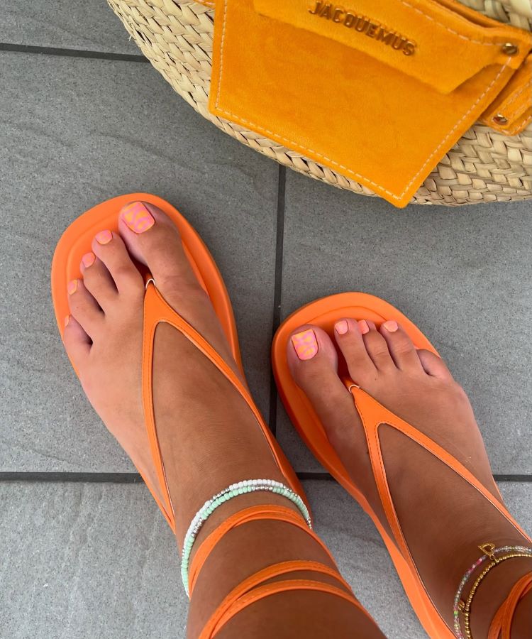 Pés com unhas decoradas em esmalte laranja neon com detalhes quadriculados. Sandálias de tiras laranjas complementam as tornozeleiras coloridas. Ao fundo, bolsa de palha com bolso amarelo da marca Jacquemus, sobre piso cinza claro. Estilo vibrante e moderno.