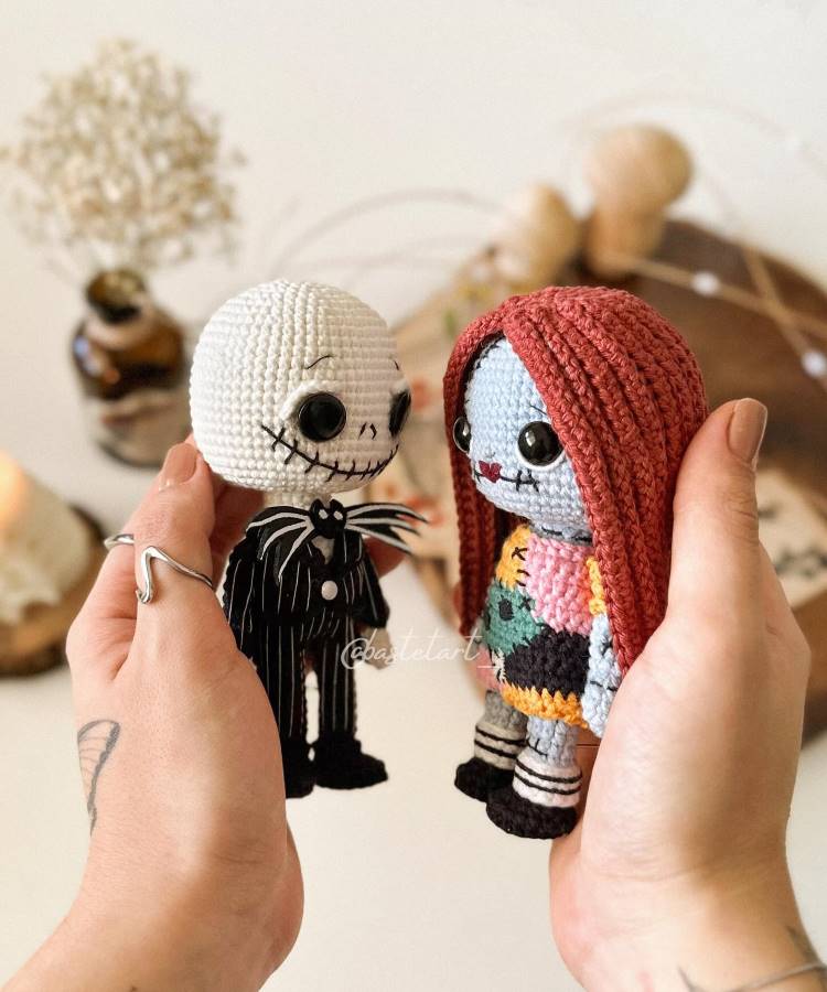 A imagem mostra duas bonecas de crochê sendo seguradas por mãos. Uma boneca é um esqueleto com terno preto e listras brancas. A outra tem cabelo vermelho e vestido colorido. Ambas têm olhos grandes e pretos. Ao fundo, há um ambiente desfocado com elementos decorativos.


