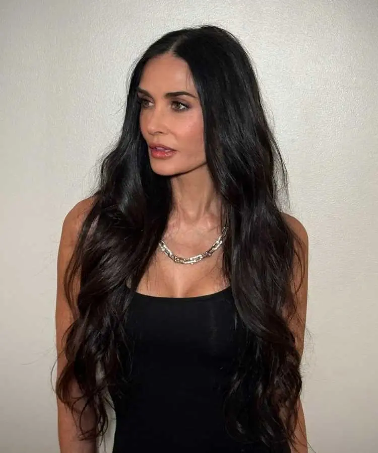 Mulher com cabelos longos, volumosos e pretos, penteados em ondas suaves. Ela veste uma blusa preta e colar prateado, com expressão serena, posicionada contra uma parede bege clara, destacando seu visual elegante e clássico.