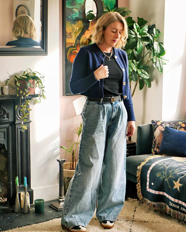 Mulher madura loira usando camiseta preta, jaquetinha curta de lã azul escuro, calça comprida que cobre os sapatos, dentro de uma casa, possivelmente uma sala com sofá, lareira e outras decorações.
