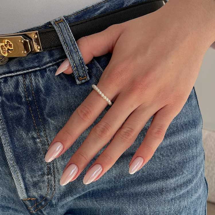 Mão feminina com unhas longas e esmalte claro, usando um anel de pérolas delicado. A mão repousa sobre jeans azul com cinto preto e detalhe metálico dourado no passador, capturando um estilo casual e elegante.