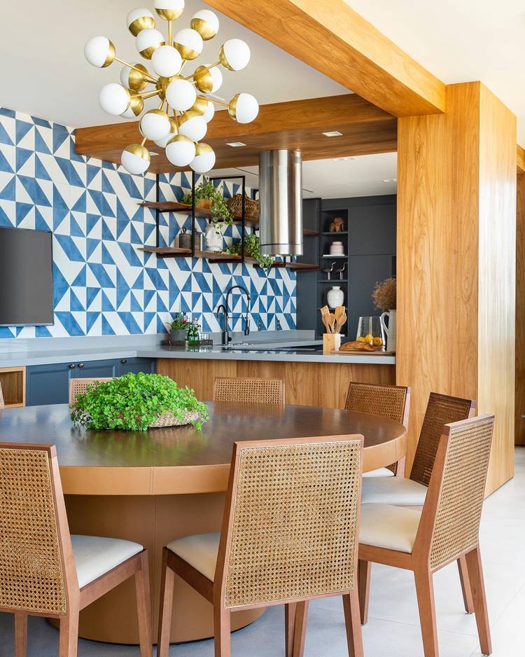 Decoração de verão de uma cozinha moderna e elegante com uma mesa de jantar. A cozinha tem um design contemporâneo, com azulejos geométricos azuis e brancos nas paredes. Há uma ilha de cozinha marrom com um tampo de mesa circular incorporado, cercado por cadeiras de madeira com encosto em treliça. Uma luminária moderna com várias lâmpadas esféricas pende do teto. Armários e prateleiras de madeira estão instalados na parede, contendo plantas e outros itens decorativos. O ambiente é iluminado e arejado, graças às cores claras e à iluminação abundante. A decoração tem um toque de verão, com cores vivas e plantas que trazem um pouco da natureza para dentro de casa.