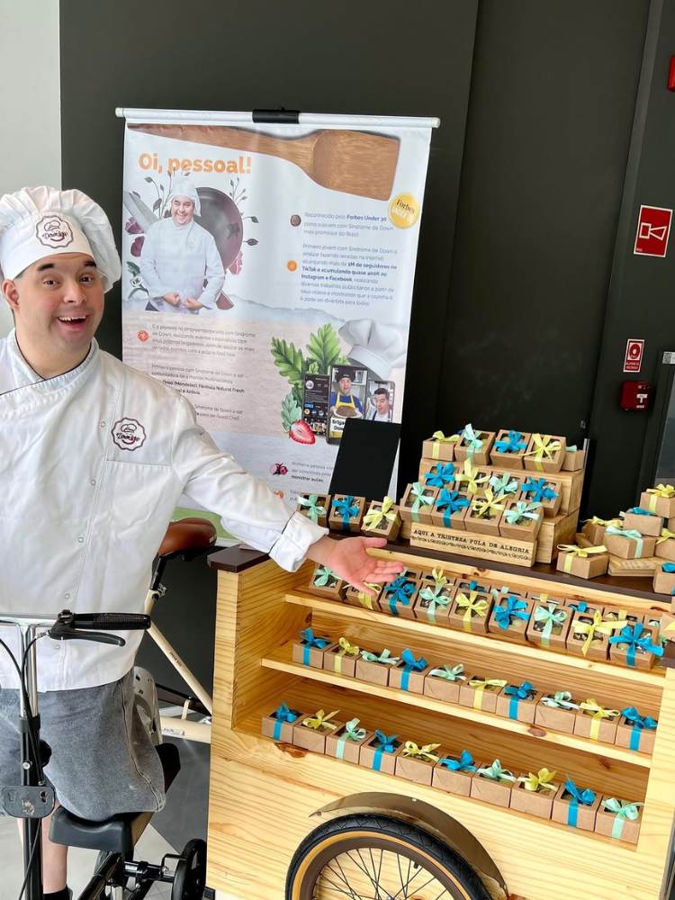 Gabriel Bernardes com roupa de cozinheiro ao lado de estande com doces