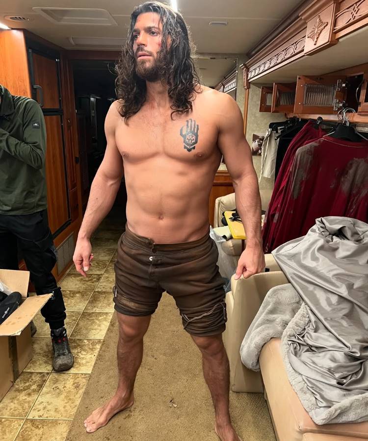Can Yaman, Homem de cabelos longos e barba, sem camisa, exibindo um físico definido e uma tatuagem no peito. Ele usa shorts marrons e está em um ambiente interno, possivelmente um trailer, com roupas e objetos espalhados ao fundo.