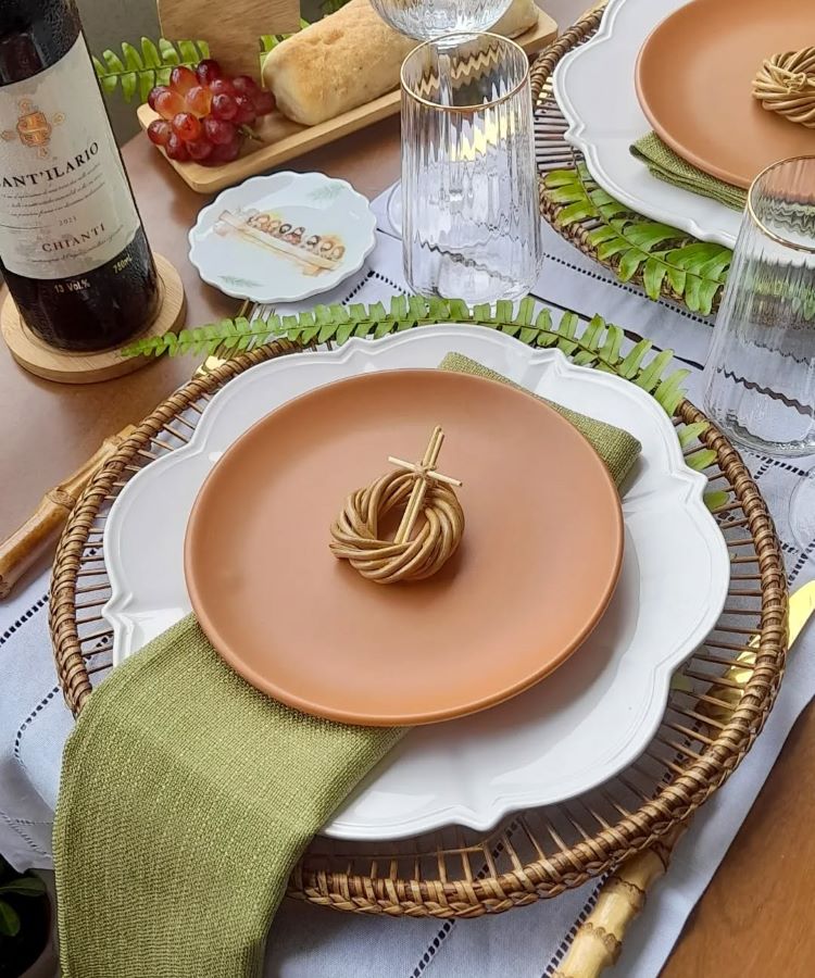 Mesa posta com inspiração rústica: sousplat de rattan, prato terracota sobre prato branco, guardanapo verde, anel decorativo de palha, taças de vidro, vinho com rótulo europeu, pão, uvas vermelhas e louça decorativa com ilustração religiosa.