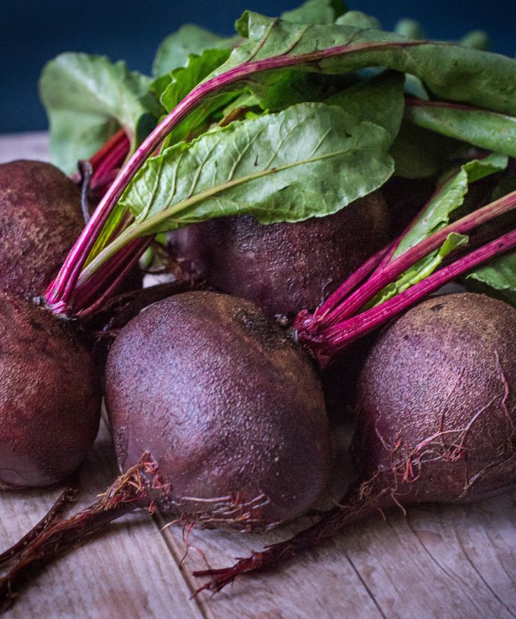 Um feixe de beterrabas frescas com as folhas verdes escuras e os caules magenta ainda intactos, dispostos sobre uma superfície de madeira clara com um fundo escuro e embaçado.