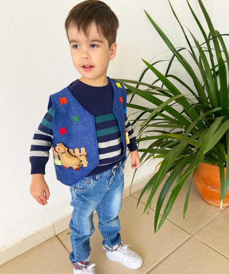 Menino com colete junino em  azul com bandeirinhas, suéter azul e calça jeans