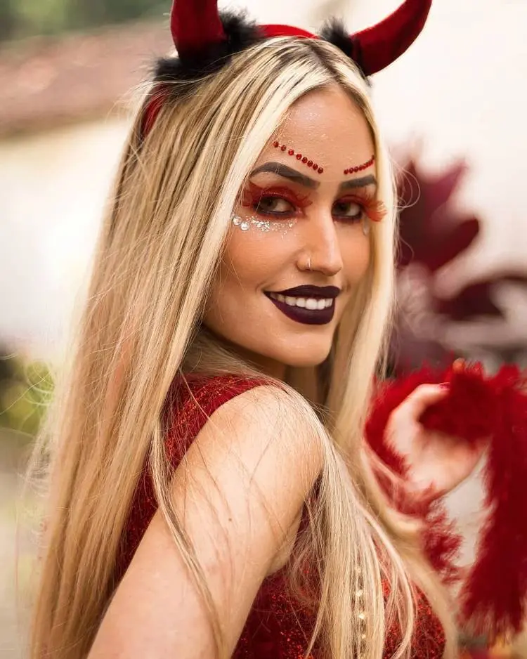 Mulher loira de pele clara usando sombra vermelha, strass vermelho e prata, batom vinho, coroa de diabo e roupa vermelha