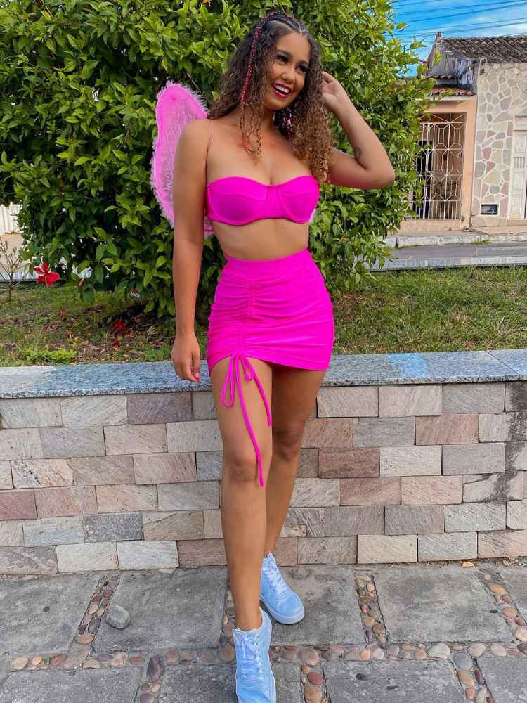 Mulher de pele morena com asa rosa de fada, cropped e saia rosas e tênis. 