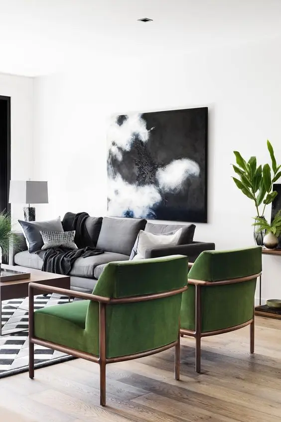 Sala de parede branca com quadro escuro, plantas, sofá cinza, tapete preto e branco, mesa de centro de madeira e poltronas verdes