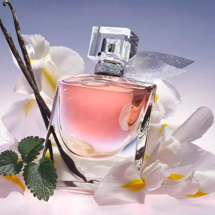 Um frasco de perfume rosado e multifacetado, "La Vie Est Belle", é o centro de uma composição floral. Rodeado por orquídeas brancas e ramos de baunilha, transmite uma sensação de feminilidade e doçura - perfumes femininos amadeirados.
