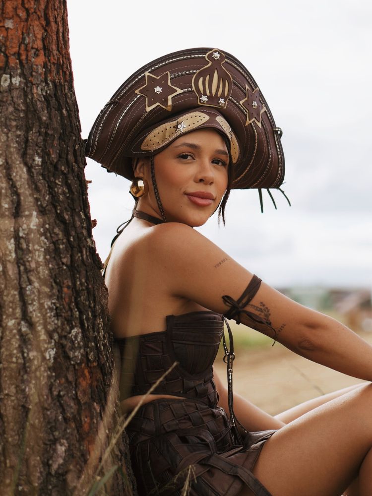 Laura Brito de roupa marrom e chapéu de cangaceiro ao lado de árvore