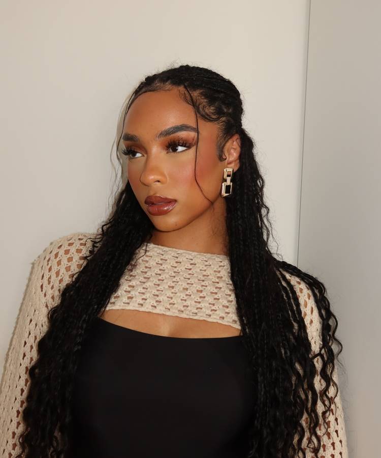 Mulher com cabelo trançado longo, baby hairs estilizados na testa. Ela usa brincos dourados geométricos, maquiagem sofisticada com batom marrom e um top preto por baixo de uma peça de crochê bege, posando contra um fundo claro minimalista.





