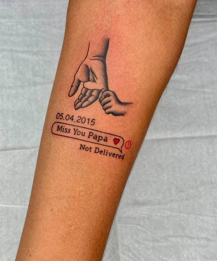 A imagem exibe uma tatuagem no antebraço de uma pessoa, apresentando uma mão adulta segurando a mão de uma criança. Abaixo, há a data "05.04.2015" e uma caixa de diálogo de mensagem com "Miss You Papa ❤️‍🩹" e "Not Delivered" em texto menor. A tatuagem é em preto e cinza.








