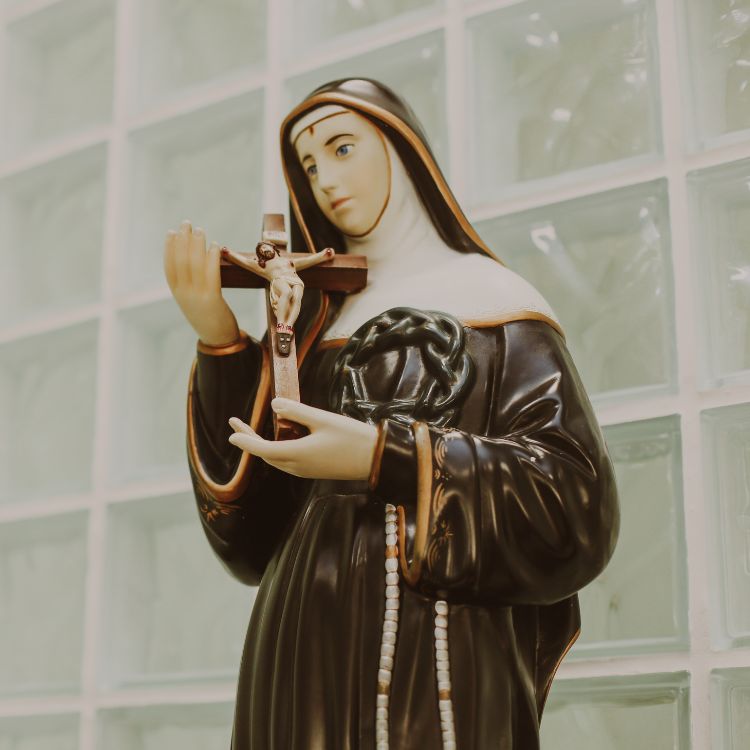 Estátua de Santa Rita de Cássia, vestida com hábito negro, segurando um crucifixo, destacando sua expressão serena, com fundo de vidros texturizados.