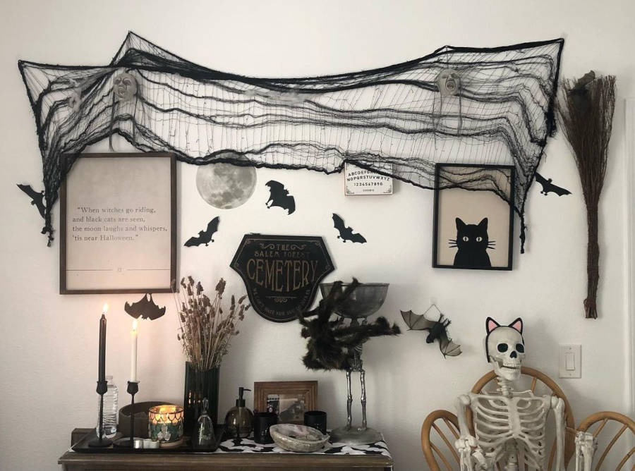 Parede branca com enfeites de halloween pretos, como quadro de gato preto, morcegos de papel, castiçal, teia, e mais