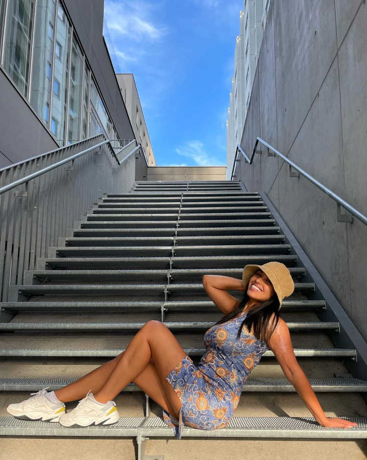 Mulher de pele morena em escada usando bucket hat bege, tênis branco e vestido azul curto