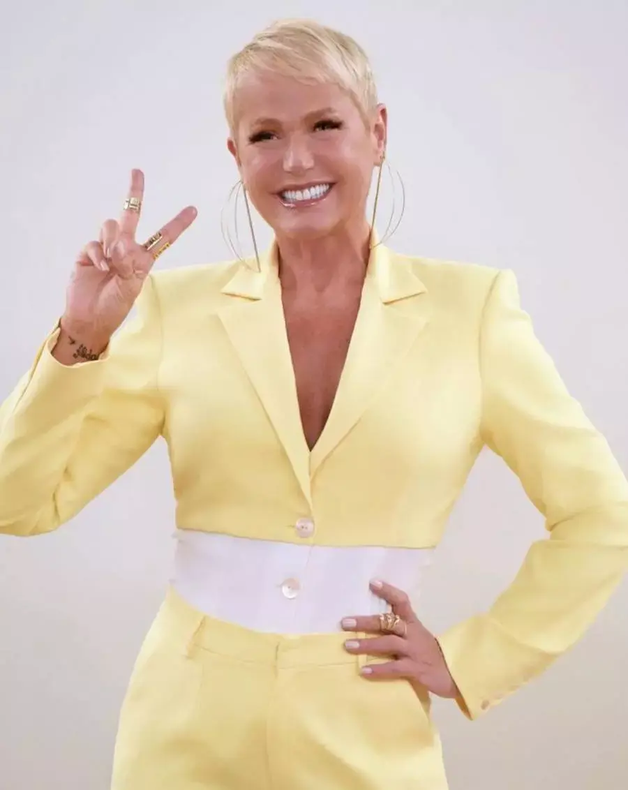 Foto de Xuxa Meneghel com um macacão amarelo com a cintura branca. A apresentadora está fazendo o sinal de paz e amor com a mão