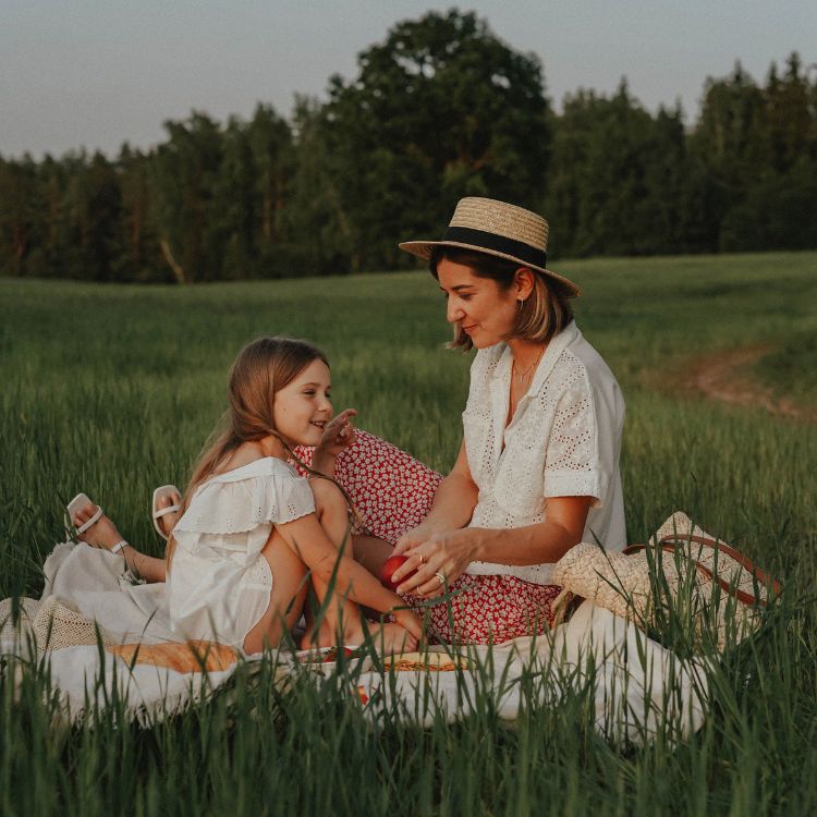 Uma mulher e uma menina compartilham um piquenique em meio a um gramado verde, cercado de árvores ao fundo. A mulher usa um chapéu de palha, enquanto ambas sorriem, criando um momento acolhedor e cheio de cumplicidade.