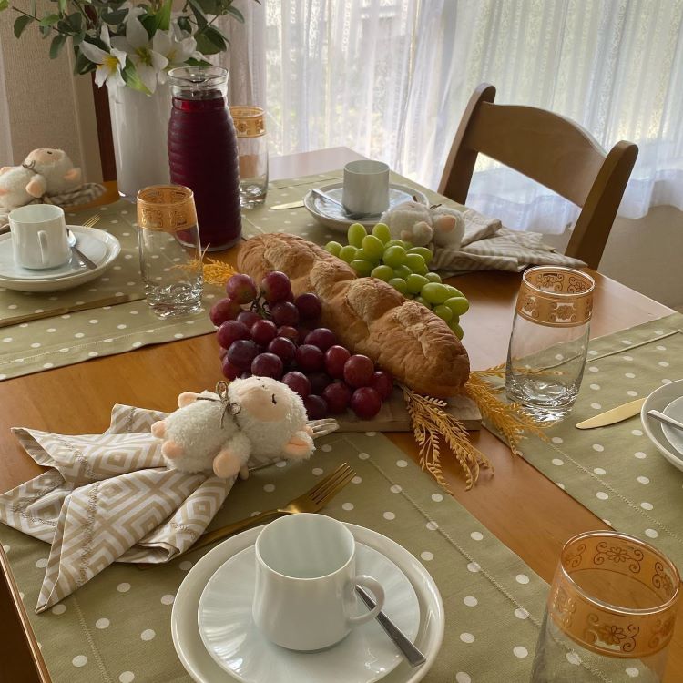 Mesa decorada com temática delicada e acolhedora: toalha verde com bolinhas brancas, pratos brancos, xícaras, talheres dourados, copos com detalhes dourados, pães, uvas, trigo seco, garrafa com suco e enfeites de carneirinhos, criando atmosfera aconchegante e festiva.