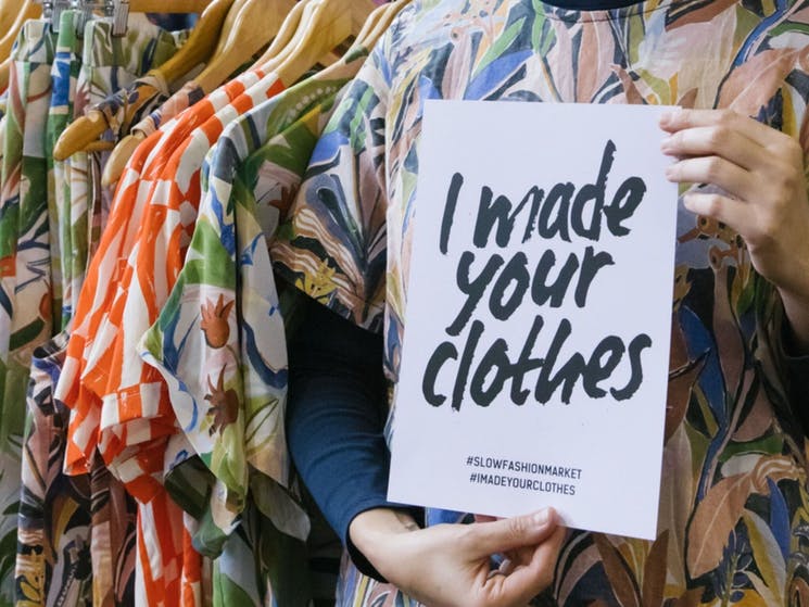 Foto de mulher na frente de arara com roupas penduradas segurando um cartaz com os dizeres: Eu faço suas roupas em inglês
