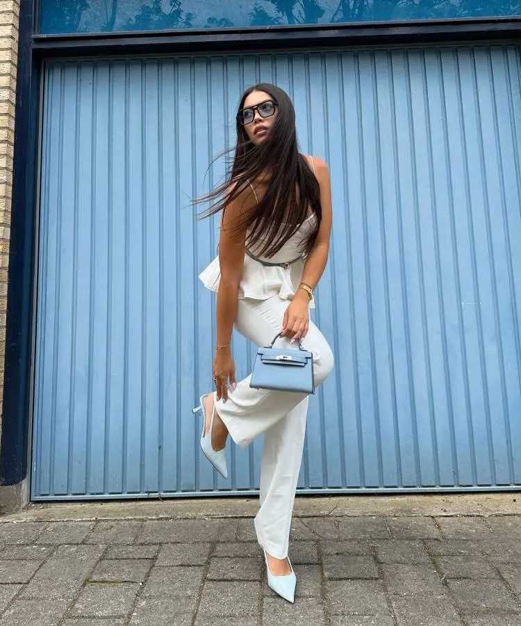 Uma mulher posa na frente de uma porta de garagem azul. Ela veste um conjunto branco, óculos de sol e sapatos de salto branco. Ela segura uma pequena bolsa azul, combinando com os sapatos e o fundo.