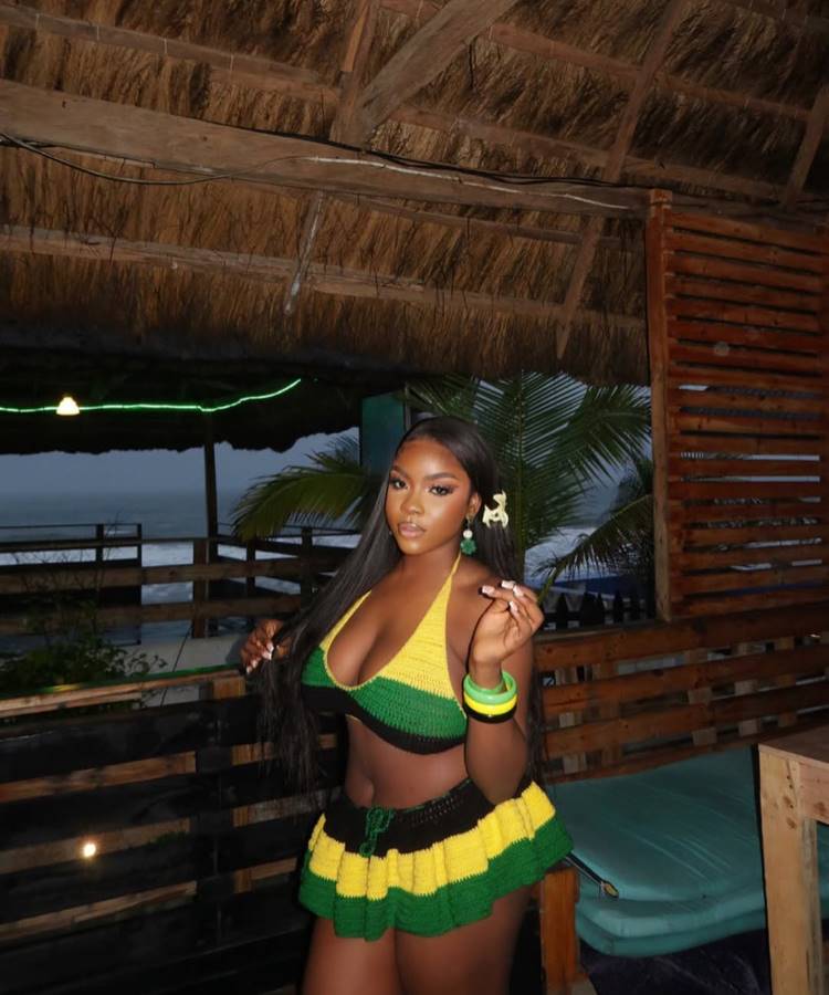 Mulher com cabelos pretos e longos posa em um bar à beira-mar, usando uma saída de praia de crochê com shorts e top. As peças são listradas em preto, verde e amarelo.