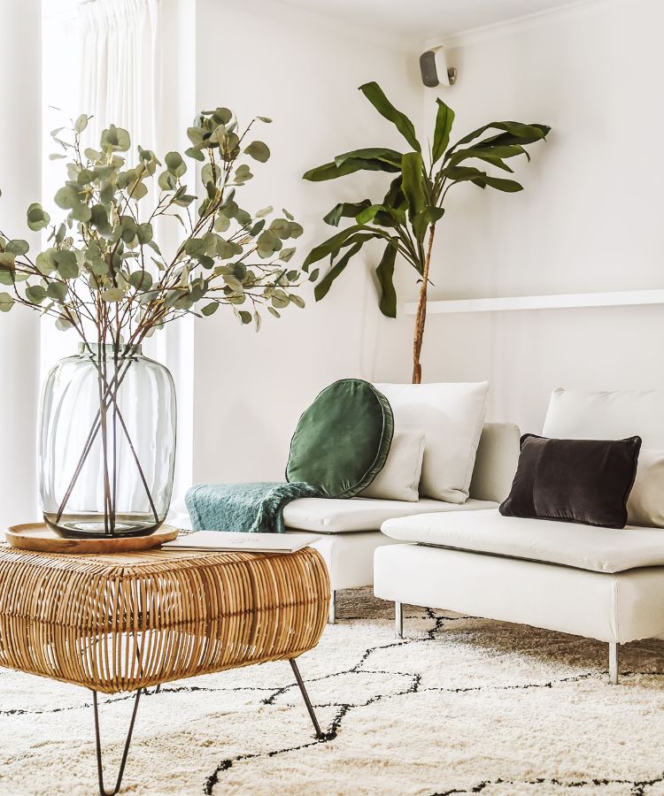 A sala é clara e arejada, com paredes brancas que refletem a luz natural. Há duas poltronas confortáveis adornadas com almofadas quadradas brancas e uma preta. Uma manta verde está casualmente jogada sobre o canto do sofá. Ao lado do sofá, há uma mesa lateral de vime com tampo circular que sustenta um grande vaso transparente. O vaso contém galhos altos com folhas verdes arredondadas, proporcionando um toque natural ao espaço. Acima do sofá na parede, há uma planta tropical pendurada que se estende para baixo em direção ao assento. O chão é coberto por um tapete texturizado em tons claros. 