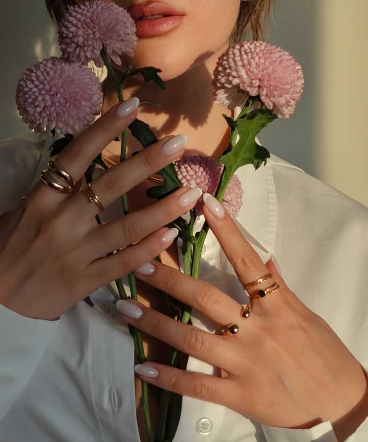 Mulher segurando flores rosa claras com hastes verdes, usando anéis dourados em dedos delicados com unhas pintadas em esmalte branco. Ela veste camisa branca e o fundo destaca luz suave, criando um contraste elegante e sereno na composição.