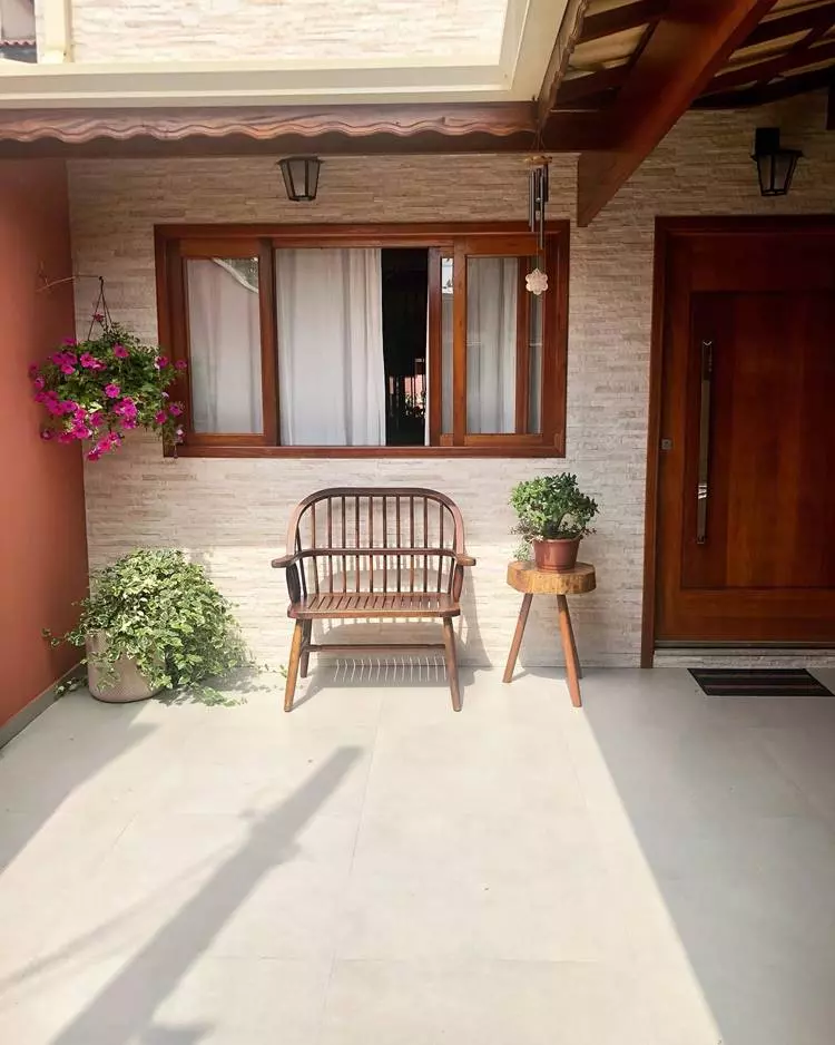 Imagem de fachada de casa simples, parede em piso de pedrinhas, janela e porta de madeira. Plantas em vasos e pendurada. Luminárias. Banco de madeira abaixo da janela.