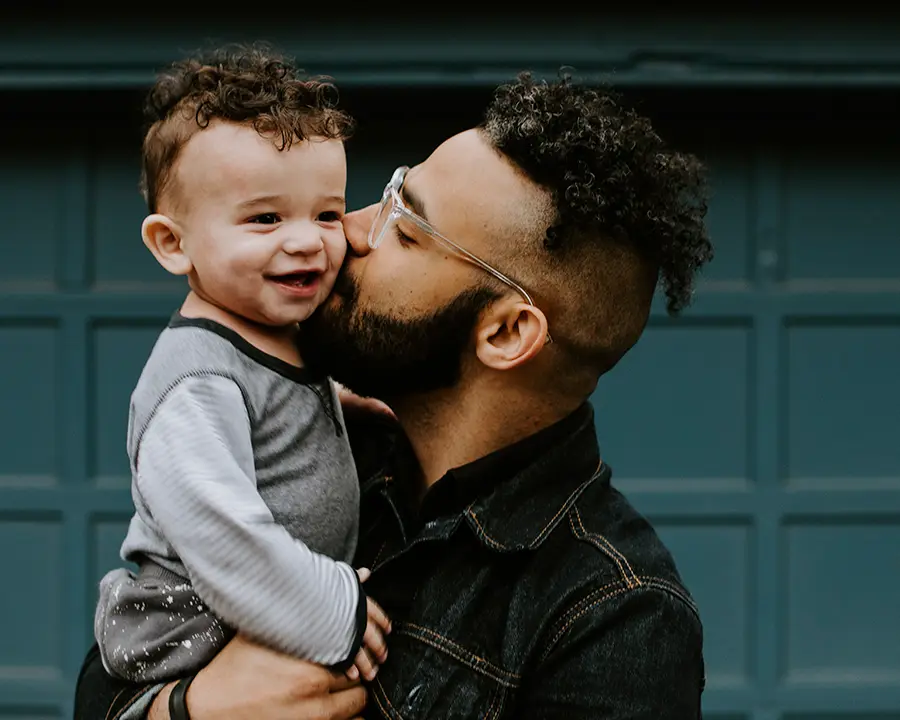 Pai de pele morena usando jaqueta jeans com filho pequeno no colo e dando um beijo na bochecha dele