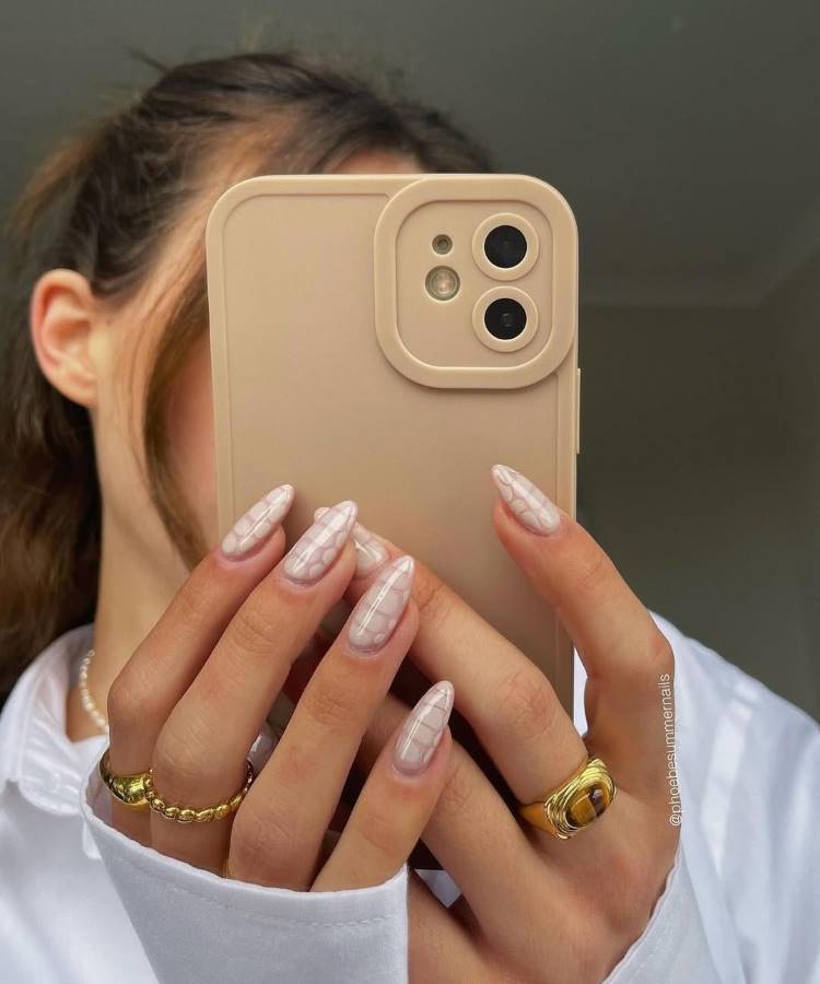 A imagem captura mãos segurando um celular bege, com unhas longas decoradas com um design xadrez branco delicado sobre base nude. Anéis dourados elegantes adornam os dedos, enquanto uma camisa branca e cabelo preso completam o visual moderno.