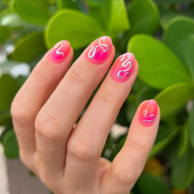 Mão exibindo estilos de unhas curtas, decoradas com esmalte rosa vibrante em degradê e detalhes ondulados brancos desenhados delicadamente. Fundo com folhas verdes iluminadas, destacando o contraste natural e realçando o visual moderno, criativo e elegante das unhas.