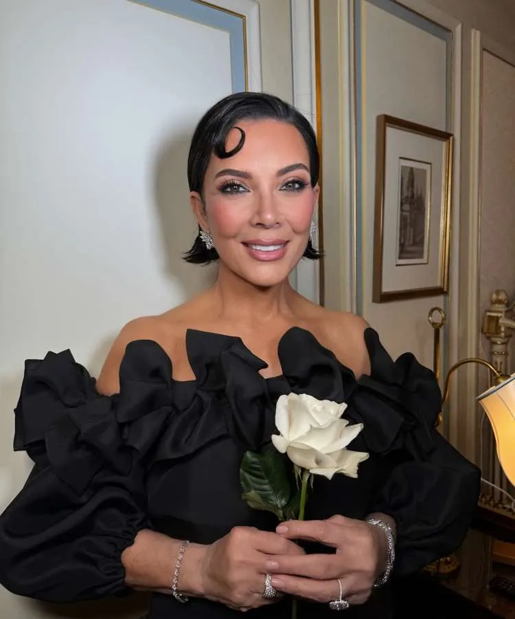 Kris Jenner, com um sorriso, segura uma rosa branca. Ela usa um elegante vestido preto com detalhes volumosos nos ombros e joias brilhantes, exibindo um penteado com uma onda lateral.