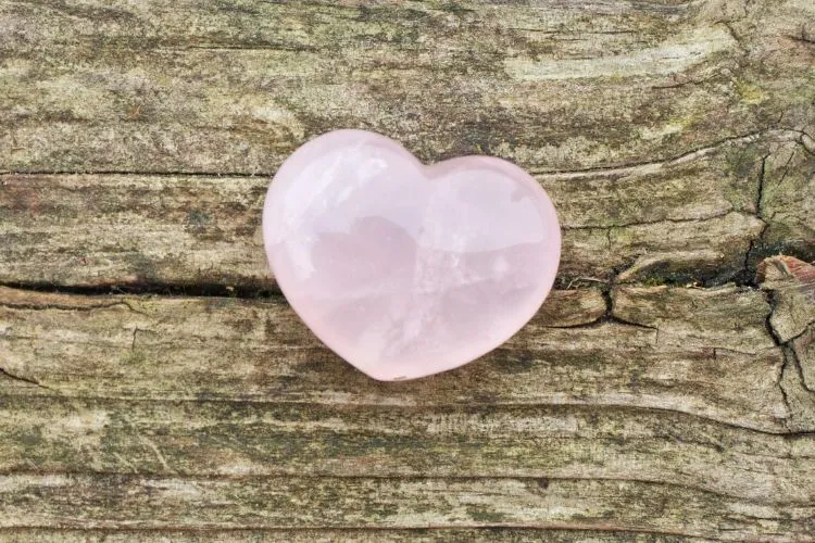 Um coração de quartzo rosa polido repousa sobre uma superfície de madeira rústica e texturizada. A pedra exibe tons suaves de rosa e reflexos da luz, enquanto a madeira ao fundo apresenta sulcos e ranhuras naturais.