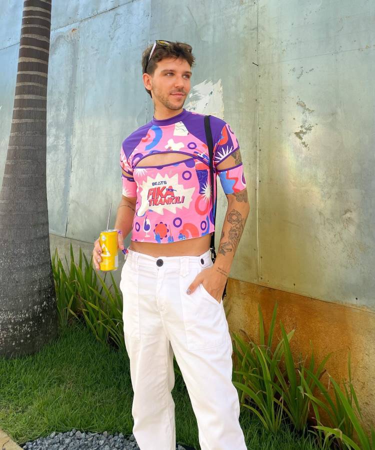 A imagem mostra um homem vestindo um abadá customizado curto, em tons de rosa, roxo e azul, com estampas tropicais e a frase "Beats Fica Tranquil". Ele combina a peça com calça branca, mochila preta e segura uma bebida.
