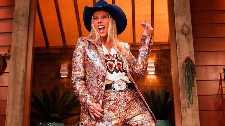 Adriane Galisteu faz pose de rockstar, e de chapéu contry e cinto de fivela faz pose como se estivesse segurando uma guitarra.