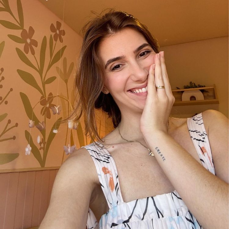 Mulher sorrindo, cabelo castanho preso, veste roupa estampada clara. Ela tem tatuagem com a palavra "CREATE" no braço. Fundo com parede decorada por flores estilizadas e tons suaves, criando ambiente alegre. A mulher posa segurando o rosto.






