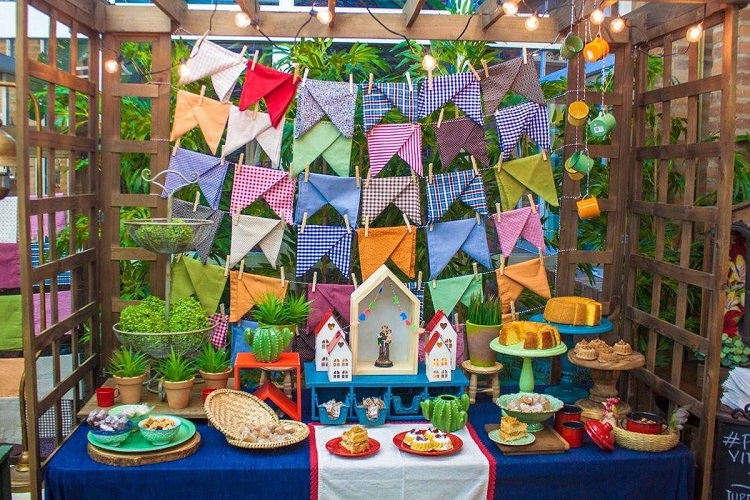A imagem representa uma decoração de festa junina colorida ao ar livre. Contém mesa com comidas como bolo e doces, além de enfeites como bandeirinha de tecido e cactos 