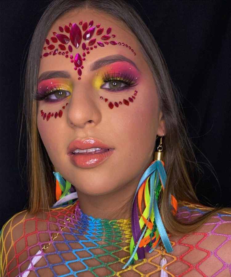Mulher de pele clara com sombra amarela, rosa e roxa, com pedrarias em vermelho na testa e abaixo dos olhos