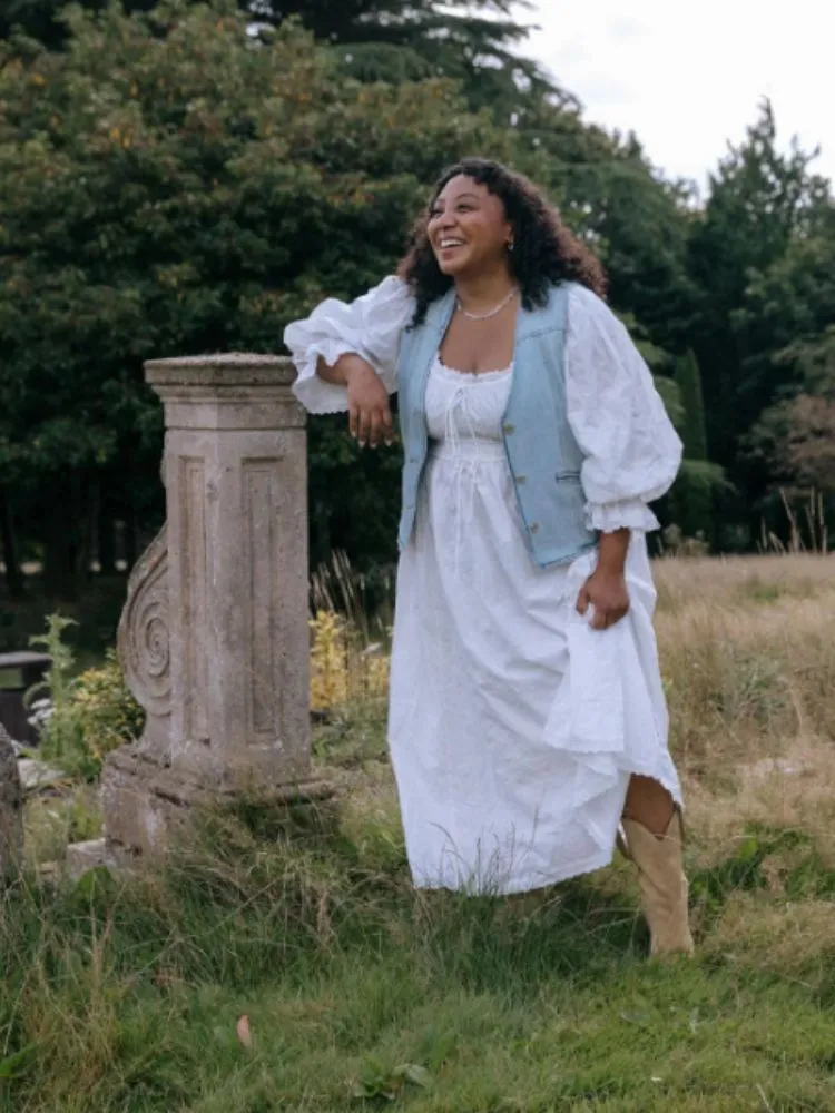 Mulher de pele negra com vestido branco longo, fluido, de mangas bufantes e colete jeans