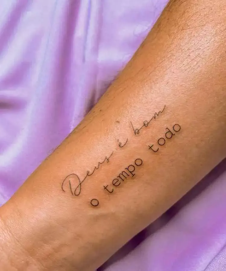 A imagem mostra uma tatuagem delicada no antebraço com a frase "Deus é bom o tempo todo". O texto combina fontes cursiva e simples, com linhas finas e elegantes. O fundo tem tecido lilás, destacando a tatuagem minimalista.
