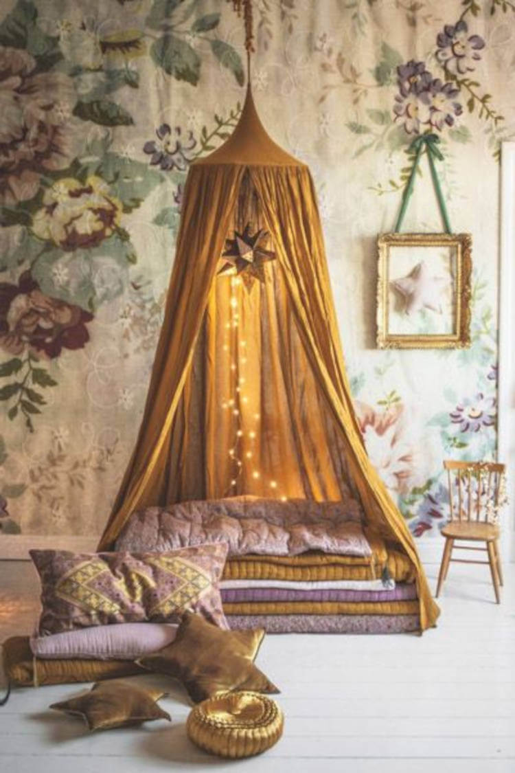 Espaço relaxante com cabana dourada, fios com lâmpadas, papel de parede florido, quadro com estrela, cadeira de madeira e almofadas e cobertas coloridas