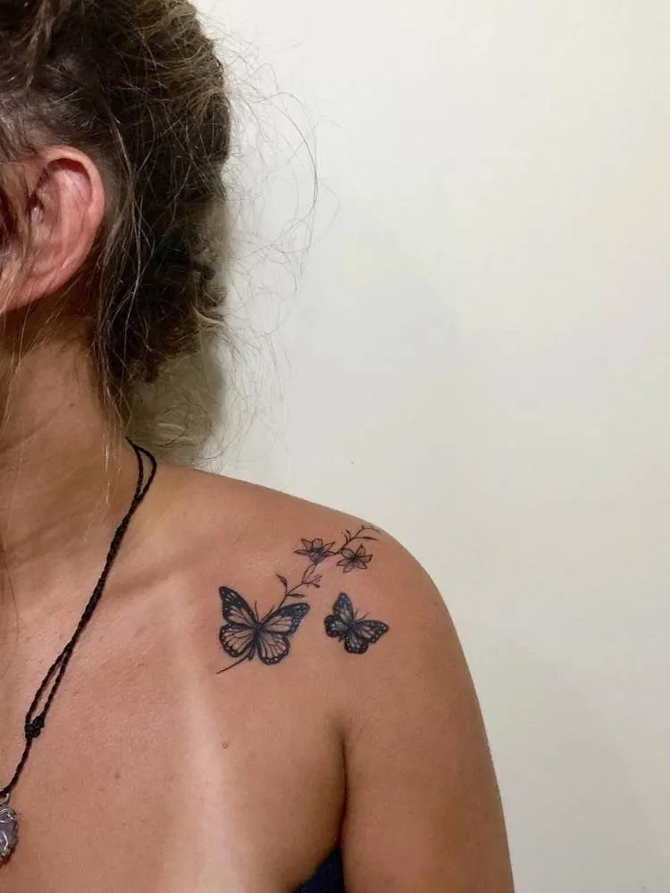 Borboletas pequenas em traços finos e um pouco mais grossos com ramos de flores no ombro de mulher de pele bronzeada, com marca da parte superior do biquíni e cabelo marrom preso.