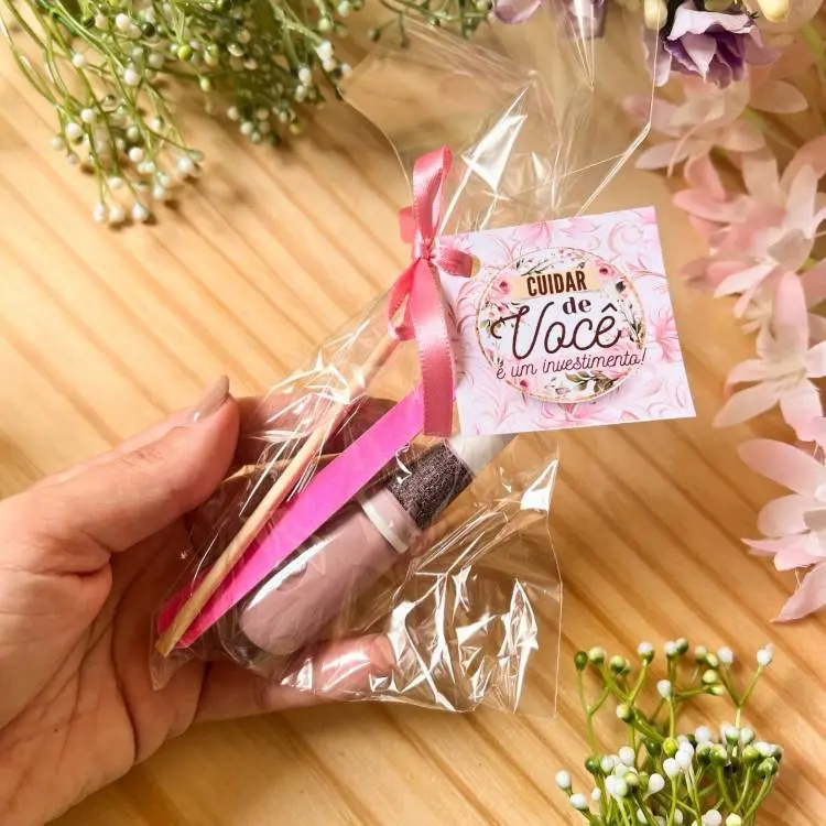 Kit delicado embalado em plástico transparente com laço rosa, contendo vidrinho de esmalte rosa, lixa de unha e palitos de madeira. Acompanhado de tag decorativa com a frase "Cuidar de você é um investimento". Fundo com flores e mesa de madeira.
