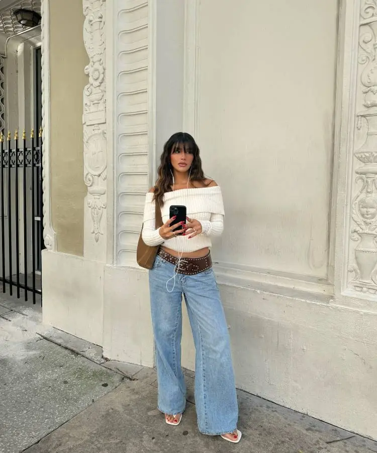 Uma mulher jovem, de cabelos castanhos ondulados, posa em frente a uma parede branca com detalhes ornamentados. Ela usa um suéter branco de ombro a ombro, calça jeans larga, cinto marrom com detalhes em metal e sandálias brancas de salto.
