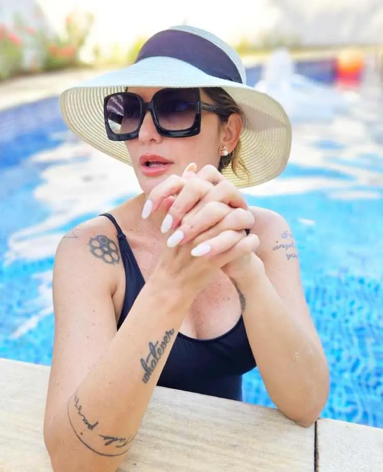 Antonia Fontenelle na piscina com maio preto, óculos escuro e chapéu, exibindo tatuagens nos pulsos, ombros e braços 